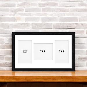 May include: A black multi-aperture picture frame with three openings, each labeled "7X5". The frame is displayed on a wooden surface against a white brick wall background. The frame is designed to hold three 17.78 x 12.7 cm photos.