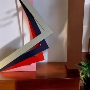 May include: Four picture frames stacked on top of each other. The frames are made of wood and are painted in different colours: silver, navy blue, red, and pink.