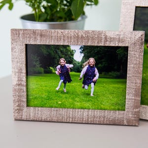 May include: Two wooden picture frames with photos of children. The frames have a distressed, light brown finish. One frame displays two girls running on a grassy lawn. The other frame shows two girls standing on the grass. A plant in a metal pot is in the background.