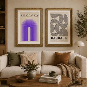 May include: Two framed Bauhaus art prints hang above a cream-colored sofa. One print features a purple arch design, the other a black and white geometric pattern. The prints are in ornate gold frames, adding a touch of elegance to the living room.