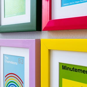 May include: A collection of framed art prints with vibrant, colorful frames. The frames are in various colors, including red, yellow, green, and blue. Some prints feature text and graphic designs, while others have abstract patterns. The artwork is displayed on a neutral wall.