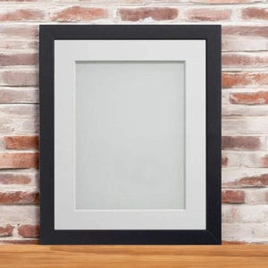May include: A black picture frame with a white mat and a white background. The frame is sitting on a wooden surface.