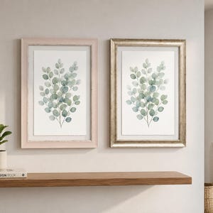 May include: Two framed botanical prints with watercolour eucalyptus leaves in green and blue hues. One frame is pale pink, the other is gold. The prints are displayed on a white wall above a wooden shelf.