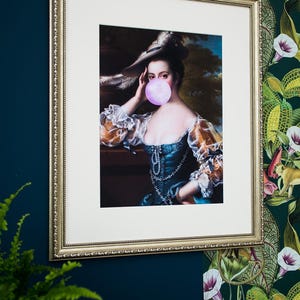 May include: Framed print of a classical portrait of a woman blowing a bubblegum bubble. The artwork is in a gold ornate frame and hangs on a dark blue wall. The woman in the portrait wears a blue dress and a hat.