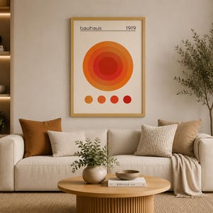 May include: A framed Bauhaus art print with a concentric circle design in shades of orange and red, with the text "Bauhaus 1919". The artwork hangs above a beige sofa with decorative pillows and a wooden coffee table. A modern living room setting.