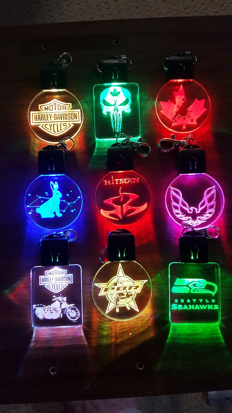 May include: Eight different keychains with various designs and colors. The keychains are made of clear acrylic and have a light inside that illuminates the design. The designs include a Harley Davidson logo, a skull, a Canadian maple leaf, a rabbit, a Hitman logo, a phoenix, a bull riding a star, and a Seattle Seahawks logo.