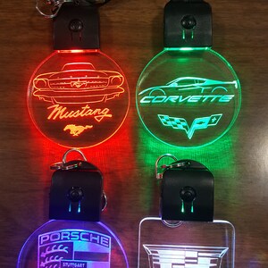 May include: Four different car logo keychains with LED lights. The keychains are round and feature the logos for Mustang, Corvette, Porsche, and Cadillac. The lights are red, green, blue, and white.