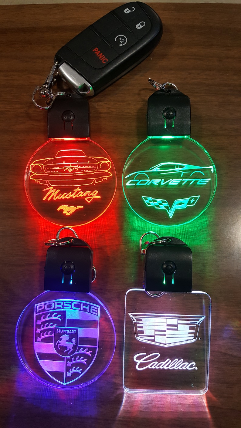 May include: Four different car logo keychains with LED lights. The keychains are round and feature the logos for Mustang, Corvette, Porsche, and Cadillac. The lights are red, green, blue, and white.