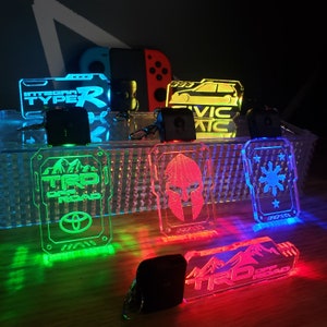 Puede incluir: Un conjunto de siete llaveros de acrílico coloridos con diferentes diseños. Los llaveros presentan una variedad de diseños, incluyendo un coche, un logotipo, una estrella y un casco. Los llaveros están iluminados con luces LED, creando una pantalla vibrante y llamativa.
