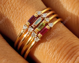 May include: A set of five gold stacking rings with colorful gemstones. The rings feature a variety of gemstones, including a red ruby, a green emerald, a blue sapphire, and a clear diamond. The rings are stacked on a finger, with the largest ring at the bottom and the smallest ring at the top.