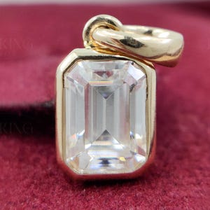 May include: A gold pendant with an emerald-cut, clear gemstone. The pendant has a rectangular shape with a gold bezel setting and a loop for a chain. The background is a deep red color.