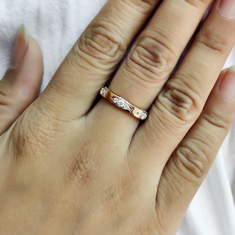 Puede incluir: Un anillo de banda en oro rosa que presenta una serie de peque&ntilde;as piedras redondas y transparentes engastadas a intervalos a lo largo de la superficie exterior. El metal tiene un acabado pulido y las piedras est&aacute;n incrustadas en la banda.