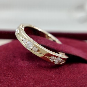 Puede incluir: Un anillo de banda dorada con una serie de piedras redondas, transparentes y facetadas engastadas a lo largo de la superficie exterior. El metal tiene un acabado pulido y reflectante, y las piedras est&aacute;n dispuestas en una fila continua.