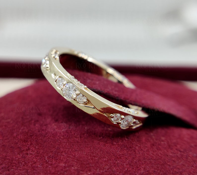 Puede incluir: Un anillo de banda dorada con una serie de piedras redondas, transparentes y facetadas engastadas a lo largo de la superficie exterior. El metal tiene un acabado pulido y reflectante, y las piedras est&aacute;n dispuestas en una fila continua.