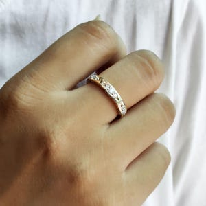 Puede incluir: Un anillo de banda dorada con una serie de peque&ntilde;as piedras claras de corte redondo engastadas en la parte superior. La banda de metal tiene un patr&oacute;n texturizado similar a un tejido que enmarca cada piedra, creando un dise&ntilde;o decorativo continuo.