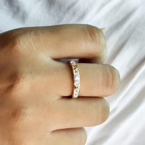 Puede incluir: Un anillo de banda de oro con una serie de peque&ntilde;as piedras claras de corte redondo engastadas en un patr&oacute;n de metal repetitivo en forma de diamante. La banda tiene un acabado pulido y se lleva en un dedo, mostrando el intrincado trabajo.