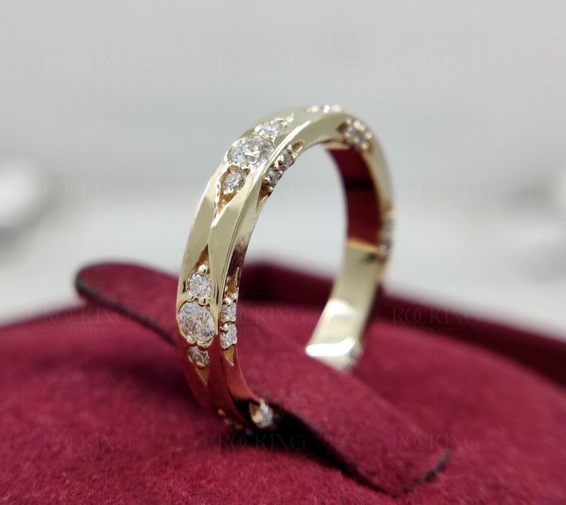 Puede incluir: Un anillo de banda de oro con una serie de piedras redondas, claras y facetadas incrustadas en la superficie met&aacute;lica. El anillo tiene un acabado pulido y un patr&oacute;n geom&eacute;trico repetitivo a lo largo de la banda, con el texto ROCKING JEWELS al fondo.