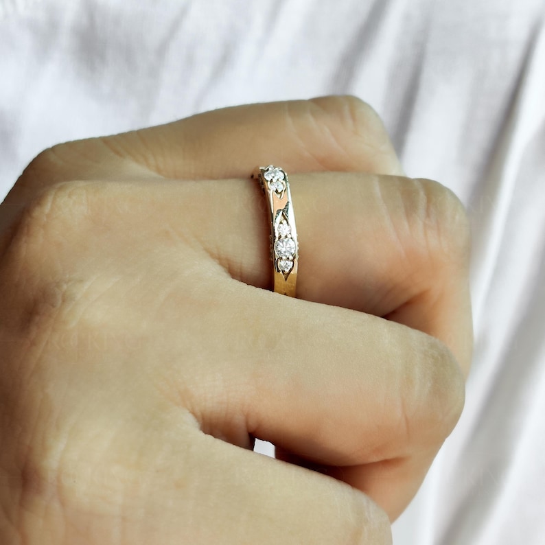 Puede incluir: Un anillo de banda dorada con una serie de peque&ntilde;as piedras transparentes de corte redondo engastadas en la superficie superior. La banda de metal tiene un acabado pulido y un perfil delgado, mostrando un patr&oacute;n decorativo que sujeta las piedras.