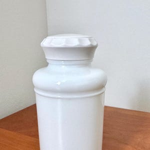 May include: A white ceramic apothecary jar with a matching lid. The jar has a cylindrical body with a slightly rounded base and a decorative band near the top and bottom. The lid is flat with a scalloped edge. The jar is sitting on a wooden surface.