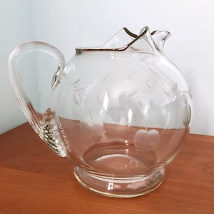 May include: A clear glass pitcher with an etched design of leaves and apples. The pitcher has a round body and a handle.