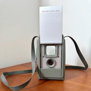 May include: A gray Anscoflex box camera featuring a tall, white, ribbed pop-up viewfinder hood with the brand name printed in red. The camera body includes a square viewfinder window, a circular lens, and a gray carrying strap attached to sides.