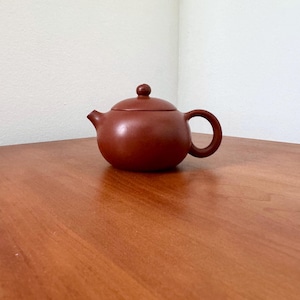 May include: A small, round, reddish-brown teapot with a matching lid and a curved handle. The teapot is made of ceramic and sits on a wooden surface. The spout is small and angled. The lid has a small knob on top.