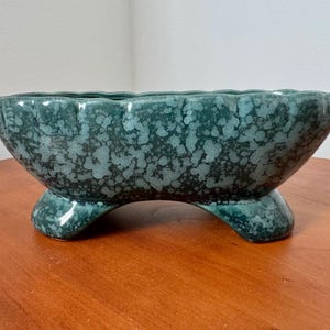 May include: A teal-colored, ceramic planter with a speckled pattern. The planter has a scalloped rim and sits on three curved feet. This decorative piece is suitable for indoor or outdoor use.