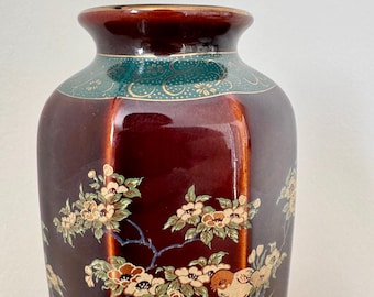 May include: A tall, glossy, maroon-colored ceramic vase with a detailed floral and bird design. The vase features a teal band with gold accents near the top and a gold rim. The design includes white, brown, and green flowers and birds.
