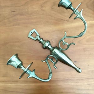 May include: Two brass candle holders with ornate, curved arms and bell-shaped cups. The candle holders have a vintage aesthetic, with decorative flourishes and a warm, metallic sheen. They are designed to be mounted on a wall.