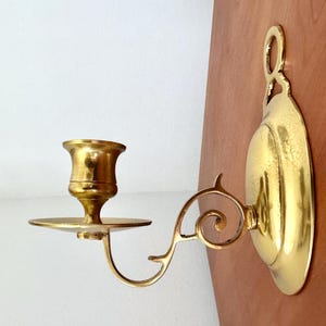May include: A brass wall-mounted candle holder. The holder features a round backplate, a curved arm with a decorative scroll, and a cup-shaped candle holder. The finish is a shiny gold color.