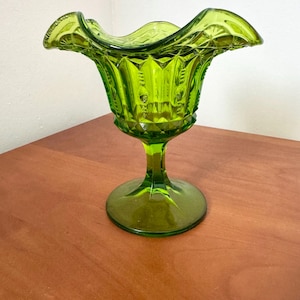 May include: A vibrant lime green glass vase featuring a pedestal base and a wide, ruffled rim. The body of the vase is decorated with a pressed diamond pattern and vertical panels, creating a textured surface that catches the light.