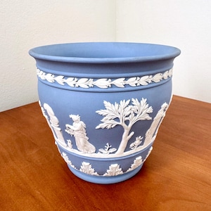 May include: A blue ceramic planter with white relief detailing. The planter features a band of leaves and classical figures around the sides. The planter is a light blue color and sits on a wooden surface.