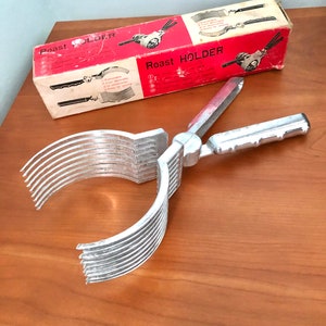May include: A silver metal roast holder with a handle and multiple prongs. The holder is designed to hold a roast while it is cooking. The holder is in its original box, which is red and white and has the words "Roast Holder" printed on it.