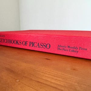 May include: A red book titled "The Sketchbooks of Picasso" with black text. The book is on a wooden surface. The book's spine includes the text "JE SUIS LE CAHIER" and the publisher's name, "Atlantic Monthly Press".