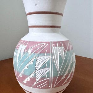 May include: A white ceramic vase with a wide base and a flared top. The vase features a pink and teal geometric design around the middle, with brown bands near the top. The vase is sitting on a wooden surface.