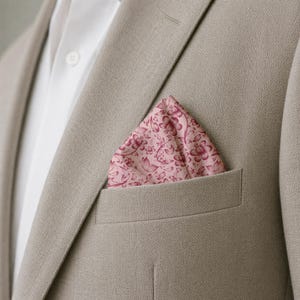 May include: A light pink pocket square with a dark pink floral pattern tucked into the breast pocket of a beige blazer. The blazer is worn over a white collared shirt with a visible button. The fabric of the blazer has a subtle herringbone texture.