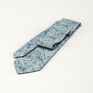 May include: A light blue necktie with a repeating floral pattern in a darker shade of blue. The tie is made of a smooth fabric and is displayed on a white background. The floral design includes stylized flowers and leaves.