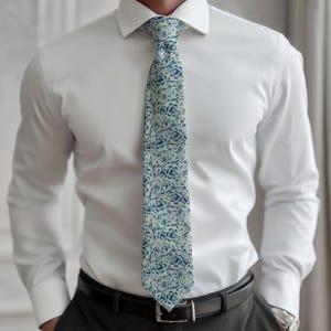 May include: A light blue tie with a repeating floral pattern in navy blue. The tie is worn over a white dress shirt and is secured with a black belt. The floral design is a classic pattern.