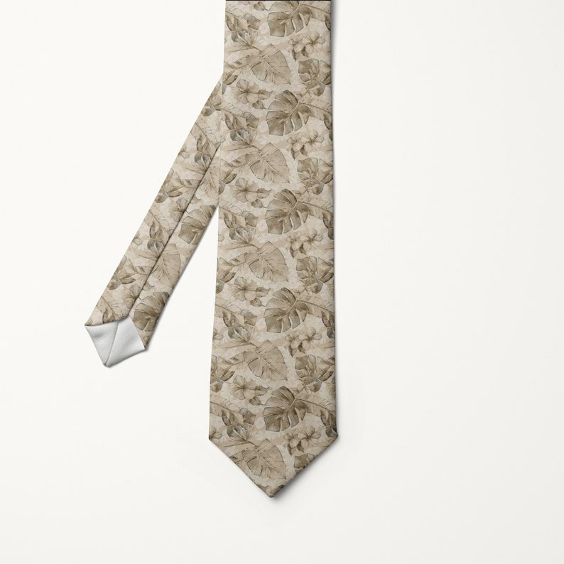 May include: A beige necktie with a repeating pattern of tropical leaves and flowers in shades of brown and tan. The tie is angled, showing the front and part of the back, against a white background.