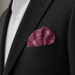May include: A dark magenta pocket square with a white tropical leaf and hibiscus flower pattern tucked into the breast pocket of a black suit jacket worn over a crisp white collared shirt with a single visible white button on the placket.
