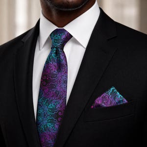 May include: A necktie and matching pocket square featuring a black base with a vibrant purple and teal mandala-inspired pattern. The set is worn with a crisp white collared shirt and a solid black suit jacket, creating a formal and coordinated look.