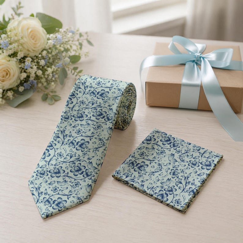 May include: A light blue floral patterned tie and matching pocket square. The tie is unrolled, displaying the full pattern. A wrapped gift box with a light blue ribbon is in the background.