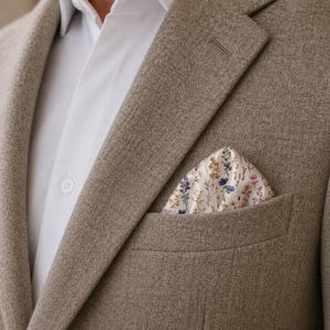May include: A tan blazer with a white dress shirt and a floral pocket square. The pocket square features a cream background with small blue, purple, and yellow flowers.