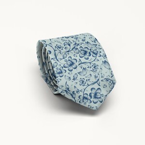 May include: Rolled necktie with a floral pattern. The tie features a light blue background with a repeating design of dark blue flowers, leaves, and vines. The tie is rolled up, showcasing the pattern and fabric.