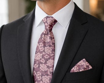 May include: A necktie and matching pocket square featuring a dusty mauve background with a detailed white floral and vine pattern. The set is worn with a crisp white collared dress shirt and a solid black suit jacket with notched lapels.