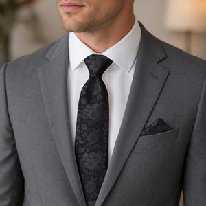 May include: A charcoal gray suit jacket worn over a crisp white dress shirt. The ensemble features a black necktie with a tonal floral pattern and a matching black floral pocket square tucked into the breast pocket of the suit jacket.