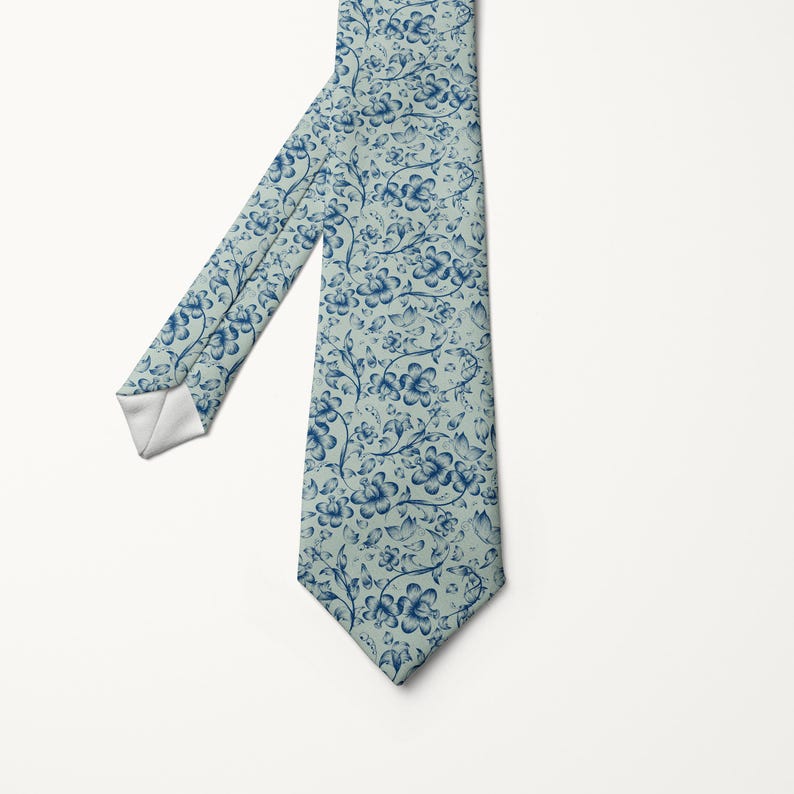 May include: A necktie with a light blue background and a repeating pattern of blue floral designs. The tie is angled, showing the front and part of the back. The tip of the tie is white.