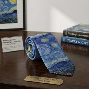 K&ouml;nnte beinhalten: Eine gerollte Krawatte mit Van Goghs Gem&auml;lde "Sternennacht" in Blau-, Gelb- und Wei&szlig;t&ouml;nen. Die Krawatte wird auf einer Holzoberfl&auml;che neben B&uuml;chern und einem Museumsausstellungsschild pr&auml;sentiert. Das Design der Krawatte ist ein ber&uuml;hmtes Kunstwerk.