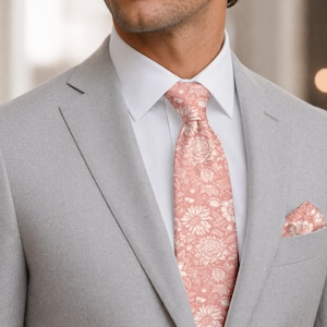 May include: A light gray suit with a white dress shirt and a floral patterned tie and pocket square. The tie and pocket square feature a pink and cream-colored floral design. The suit jacket has a notched lapel.