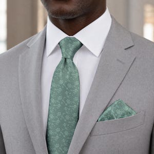 May include: A light gray suit with a white dress shirt, and a sage green tie and pocket square. The tie and pocket square feature a floral pattern. The suit jacket has a notched lapel and a single-breasted design.